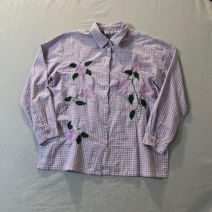 May include: A long-sleeved button-down shirt with a gingham pattern in white and lavender. The shirt features embroidered lavender flowers with green leaves. The shirt has a classic collar and buttoned cuffs.