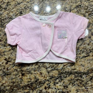 May include: A light pink short-sleeved bolero jacket with white trim along the edges. The garment features two round buttons at the neckline and a small square patch on the left chest with a floral pattern and a tiny white ribbon bow detail.