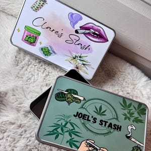 May include: Two rectangular metal stash boxes. One box has the text "Clare's Stash" with illustrations of a lighter, cannabis, and lips smoking a joint. The other box has the text "Joel's Stash" with cannabis leaves and a hand rolling a joint.