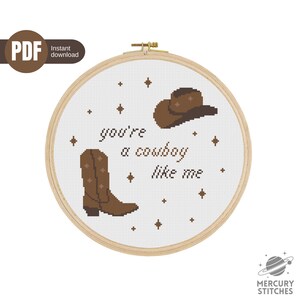 Peut inclure: Un cercle de broderie au point de croix avec un motif de botte et de chapeau de cowboy marron et le texte "you're a cowboy like me". Le motif est sur fond blanc avec des étoiles. Le cercle est en bois.