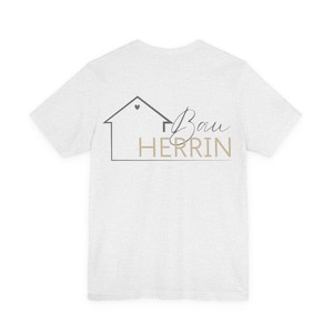 Puede incluir: Camiseta gris claro con un gráfico de una casa y el texto "Bau Herrin" en negro y marrón. El gráfico de la casa tiene un corazón en el tejado. La camiseta es de un material suave.
