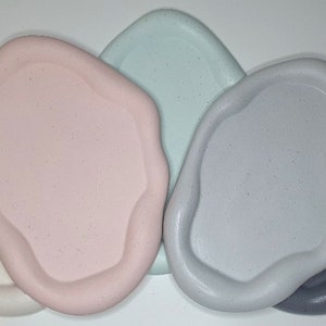 May include: Five irregularly shaped, ceramic coasters in various shades of grey, light blue, pink, and white.