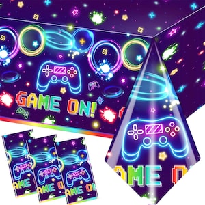 May include: A purple and black plastic tablecloth with a neon green and blue video game controller design. The tablecloth has the words "Game On!" printed on it. There are three matching napkins with the same design.