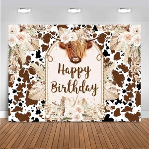 May include: A brown and white cowhide patterned backdrop with a brown highland cow head and floral accents. The backdrop has the text "Happy Birthday" in gold glitter lettering.
