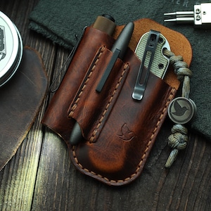 May include: A brown leather pocket organizer with a pen, knife, and flashlight. The organizer has a bird logo and is next to a green and gray lanyard. Other items include a leather balm tin, a lighter, and metal tools.