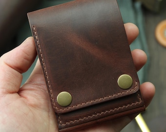 May include: A brown leather wallet with a snap closure. The wallet is rectangular and features two brass-colored snaps. The stitching is visible along the edges. The wallet is held in a hand.