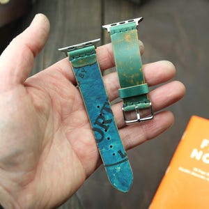 Puede incluir: Dos correas de reloj de cuero en tonos verdes y azules. Una correa tiene un aspecto envejecido y vintage con la palabra "PORT" visible. La otra correa es de color verde liso. Ambas correas tienen herrajes plateados.