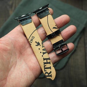 May include: Two tan leather watch bands with black hardware are held in a hand. The bands have a printed design with the words "NORTH" and "ING" visible. The bands are for a smartwatch.