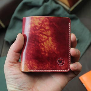 May include: A handcrafted, bi-fold leather wallet in a rich, reddish-brown hue with a unique, mottled pattern. The wallet features white stitching and a small, embossed bird logo. The wallet is held in a hand.