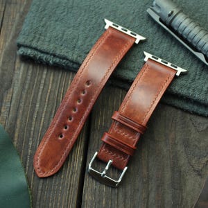Può includere: Due cinturini per orologi in pelle marrone con chiusure e hardware color argento sono esposti su una superficie di legno. I cinturini sono cuciti e hanno un colore ricco e caldo. Uno è allacciato e l'altro è slacciato, mostrando i fori di regolazione.