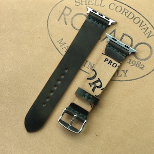 Könnte beinhalten: Zweiteiliges dunkelgrünes Leder-Uhrenarmband mit silberfarbenen Beschlägen. Das Armband wird auf einer hellbraunen Oberfläche mit einem gestempelten Logo gezeigt. Es hat eine Schnalle und mehrere Verstelllöcher.