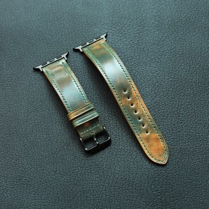 May include: Two leather watch straps in green and brown with black metal buckles. The straps are on a dark grey surface.
