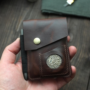 May include: A brown leather wallet with a flap closure and a brass button. The wallet has a coin pocket with a decorative metal emblem. A pen is held in place by a black elastic band on the side.