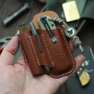 May include: A brown leather pocket organiser with a pen, knife, and flashlight. The organiser has a bird logo and is held in a hand. A lanyard with a bird logo is attached.