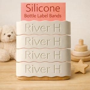May include: A white silicone bottle label band holder with the text "River H" repeated four times. The holder has a wooden base and a pink label that reads "Silicone Bottle Label Bands". A teddy bear and wooden toys are in the background.