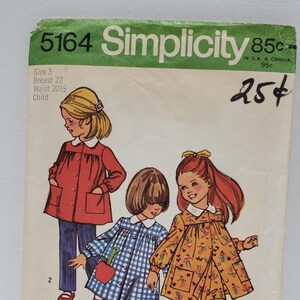 May include: A vintage Simplicity sewing pattern for children's dresses. The pattern number is 5164 and the price is 85 pence. The pattern includes three different dress styles: a red dress with pockets, a blue and white gingham dress, and a yellow floral dress.