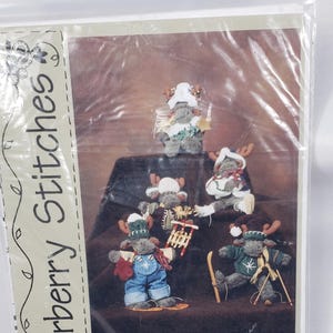 Peut inclure: Un emballage plastique transparent contenant un patron de couture intitulé "MOOSE ON THE LOOSE #T131". Le patron présente des figurines d'orignaux vêtus de tenues d'hiver, avec des chapeaux, des salopettes et des skis. Le logo Tenderberry Stitches est visible.