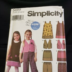 May include: Simplicity sewing pattern number 5489 for sizes 3-8. The pattern includes instructions for making a dress, waistcoat, skirt, and trousers. The pattern is illustrated with a model wearing a pink waistcoat, brown trousers, and a brown dress.