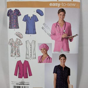 May include: Simplicity 3932 sewing pattern for scrub tops and caps in sizes AA 10-18. The envelope displays illustrations and photographs of various scrub top styles with V-necks, short sleeves, and pockets, alongside matching surgical caps.
