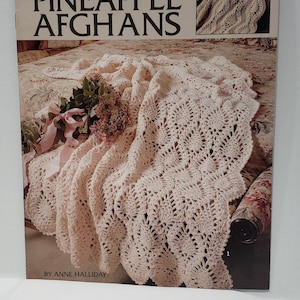 May include: A book titled "So Pretty Pineapple Afghans" by Anne Halliday. The cover features a crocheted pineapple stitch afghan draped over a chair with a bouquet of flowers on the seat. The book includes 6 crochet patterns.