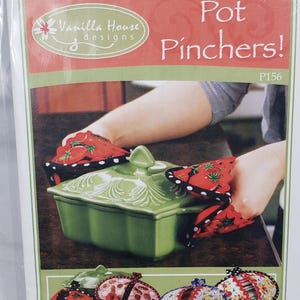Puede incluir: Un paquete con "Pot Pinchers!" con una fuente verde para hornear y agarraderas rojas con un diseño de fresa. El paquete también muestra varios diseños de agarraderas.