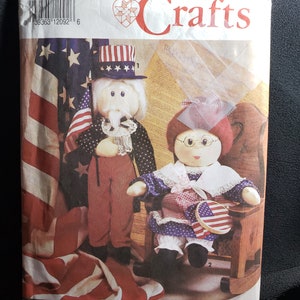 May include: Simplicity Crafts sewing pattern featuring patriotic dolls. The pattern cover displays Uncle Sam and a doll in a rocking chair, both adorned in red, white, and blue attire. The background includes an American flag and the text "Simplicity Crafts".