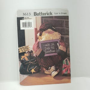 May include: A sewing pattern for a Santa Claus doll holding a chalkboard that says "...only 12. Days 'til Christmas!" The doll is wearing a red and black plaid shirt and a white beard. The pattern is from Butterick and is for one size.