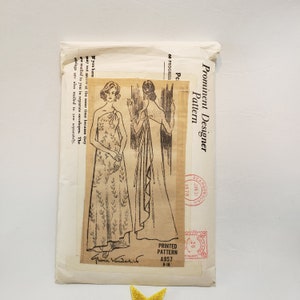 May include: A vintage sewing pattern envelope with a black and white illustration of a woman wearing a long, one-shoulder dress. The envelope is labeled "Prominent Designer Pattern" and "Printed Pattern A957 8-18".
