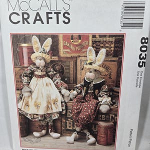 May include: McCall's Crafts pattern featuring two bunny dolls. The dolls are dressed in patterned fabric dresses and hats, with long, floppy ears. The text reads "Hop to it! These cute bunnies are 34 inches tall."
