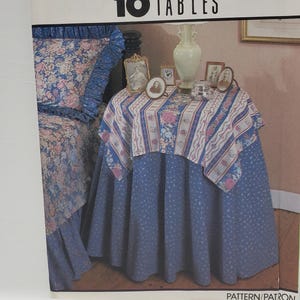 May include: Simplicity House 10 Tables sewing pattern, pattern number 8418, one size, featuring a blue and white floral tablecloth with a ruffled edge, on a blue and white floral table skirt, with a vase and framed pictures on top.