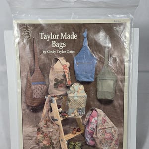 May include: A sewing pattern envelope titled Taylor Made Bags by Cindy Taylor Oates, model TA-20. The cover displays various handmade fabric bags in floral, denim, and textured patterns, featuring shoulder straps and exterior pockets on a brown background.