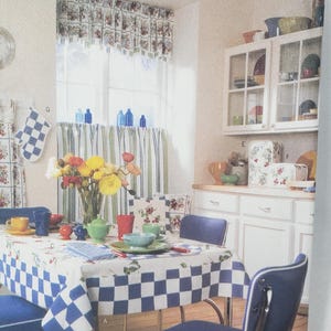May include: A kitchen scene with a blue and white checkered tablecloth and matching chairs. The room features floral curtains, a window valance, and a cabinet with dishes. A vase of flowers sits on the table.