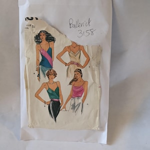 May include: A vintage sewing pattern envelope featuring illustrations of four different tops. The envelope is torn and has handwritten text, including "Butterick 3158" and "B $1.58". The tops are depicted in various colors and styles.