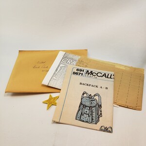 May include: A selection of sewing patterns for a backpack. The patterns include a tan envelope with "Quilted Back Pack" written on it, a McCall's pattern, and a gold glitter star.