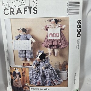 May include: McCall's Crafts pattern showcasing cow-themed plush doll designs. The pattern provides instructions for crafting different cow dolls, including one holding a toilet roll and another wearing a dress. The pattern is labelled "8590".