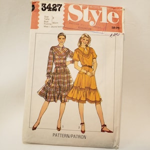 May include: Vintage sewing pattern packet featuring illustrations of two dresses. One dress is plaid with long sleeves, the other is solid orange with short sleeves. The packet has the word "Style" and the number "3427" printed on it.