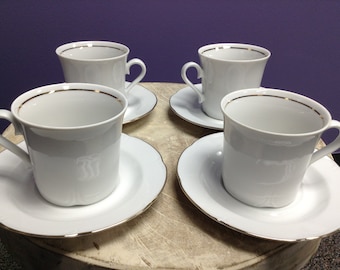 May include: Four white teacups with gold trim and matching saucers. The cups and saucers are arranged on a wooden surface.