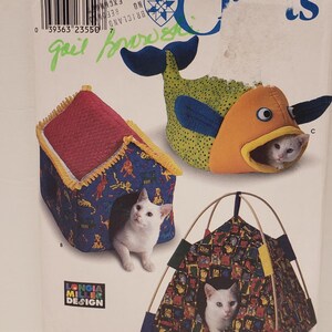 May include: A Simplicity Crafts sewing pattern for making three cat beds. The pattern includes a house-shaped bed, a fish-shaped bed, and a tent-shaped bed. Each bed features a different colorful fabric with a cat-themed design. The pattern is labeled "9004" and "One Size".