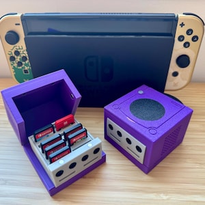 May include: A purple GameCube-style game cartridge holder and a matching purple cartridge case, both with a light gray base. The open case holds several red and black game cartridges. A Nintendo Switch console is in the background.