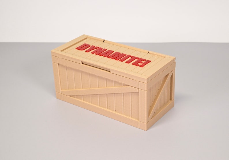 Puede incluir: Una caja de madera de color beige con una etiqueta roja que dice "&iexcl;Dinamita!" en la parte superior.