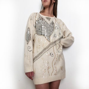 May include: A white sweater with a large silver sequin design. The sweater has a crew neck and long sleeves.