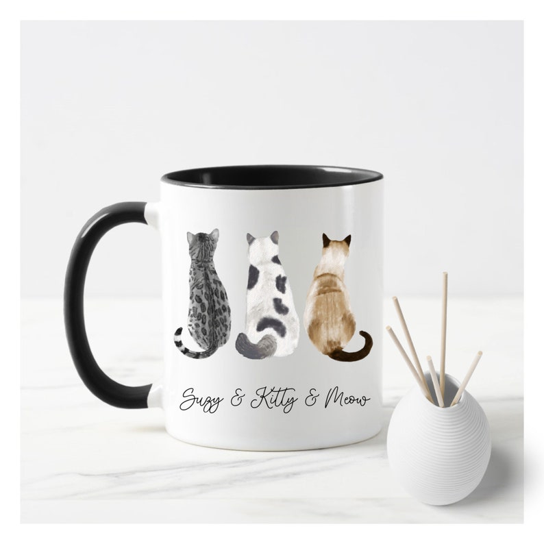 May include: White ceramic mug with a black rim and handle. The mug features a watercolor illustration of three cats sitting back to back. The text "Suzy & Kitty & Meow" is printed below the cats.