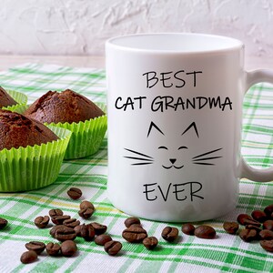 May include: White ceramic mug with black text that reads "Best Cat Grandma Ever" and a cartoon cat face. The mug is on a green and white checkered tablecloth with coffee beans and chocolate muffins.