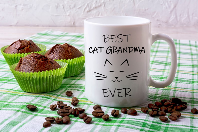 May include: White ceramic mug with black text that reads "Best Cat Grandma Ever" and a cartoon cat face. The mug is on a green and white checkered tablecloth with coffee beans and chocolate muffins.