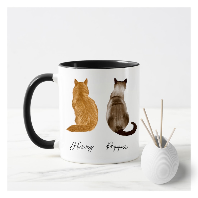 May include: White ceramic coffee mug with a black rim and handle. The mug features a watercolor illustration of two cats sitting back to back. The cat on the left is orange and the cat on the right is gray. The text "Harvey" is written below the orange cat and "Popper" is written below the gray cat.