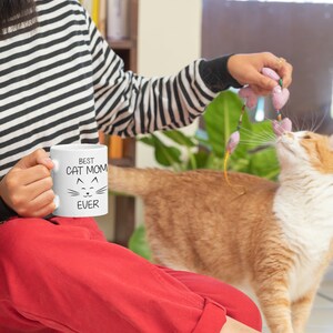May include: A woman wearing a black and white striped shirt and red pants is holding a white mug with the text "BEST CAT MOM EVER" and a cat face. She is playing with a cat with a pink and green toy.