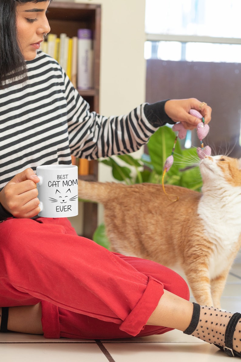 May include: A woman wearing a black and white striped shirt and red pants is holding a white mug with the text "BEST CAT MOM EVER" and a cat face. She is playing with a cat with a pink and green toy.