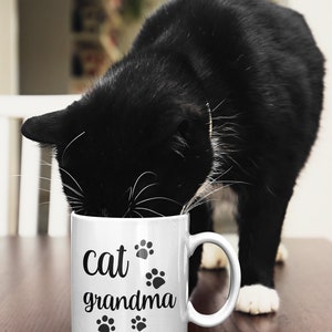 May include: White ceramic mug with black text that reads "cat grandma" and paw prints. A black and white cat is drinking from the mug.