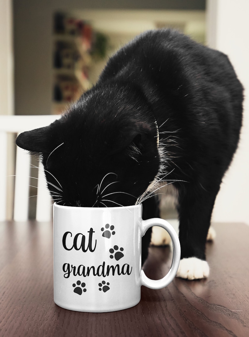 May include: White ceramic mug with black text that reads "cat grandma" and paw prints. A black and white cat is drinking from the mug.