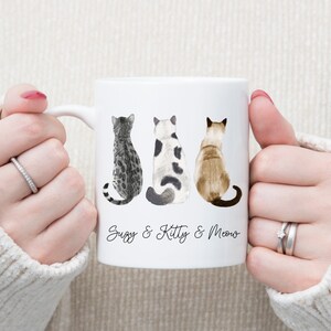 May include: White ceramic mug with three cats sitting back to back. The cats are a tabby, a black and white cat, and a Siamese. The mug has the text "Suzy & Kitty & Meow" printed on the bottom.